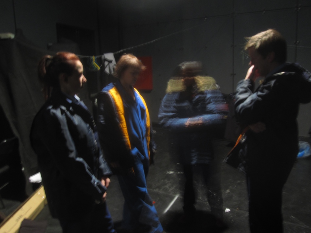 The cast in overalls and Parker coats wait pensively for their time to go on stage.