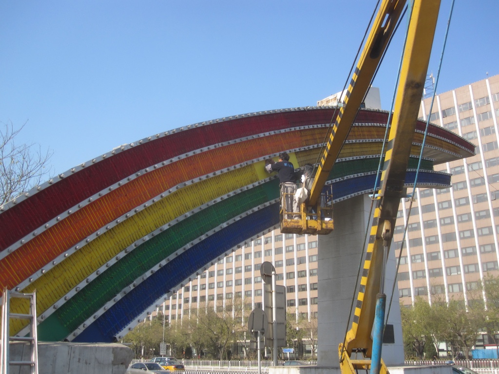 From the platform of a large yellow crane a worker replaces light bulbs on a huge rainbow sign.