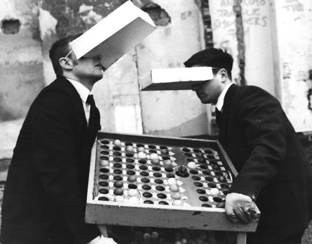 Two besuited men carry a bingo machine between them over wateground, they wear protective gloves and have blank cereal boxes over their eyes like VR goggles.