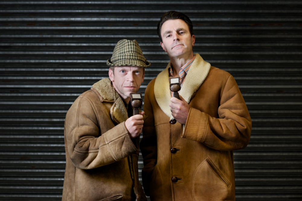 Two commentators in sheepskin coats holding lip microphones stood in front of a metal roller shutter.
