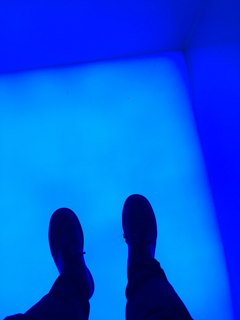 A point of view shot looking down on a glowing blue floor in the corner of a glowing blue room. The photographer's shoes and the legs from the knees down intrude on the bottom edge of the frame.