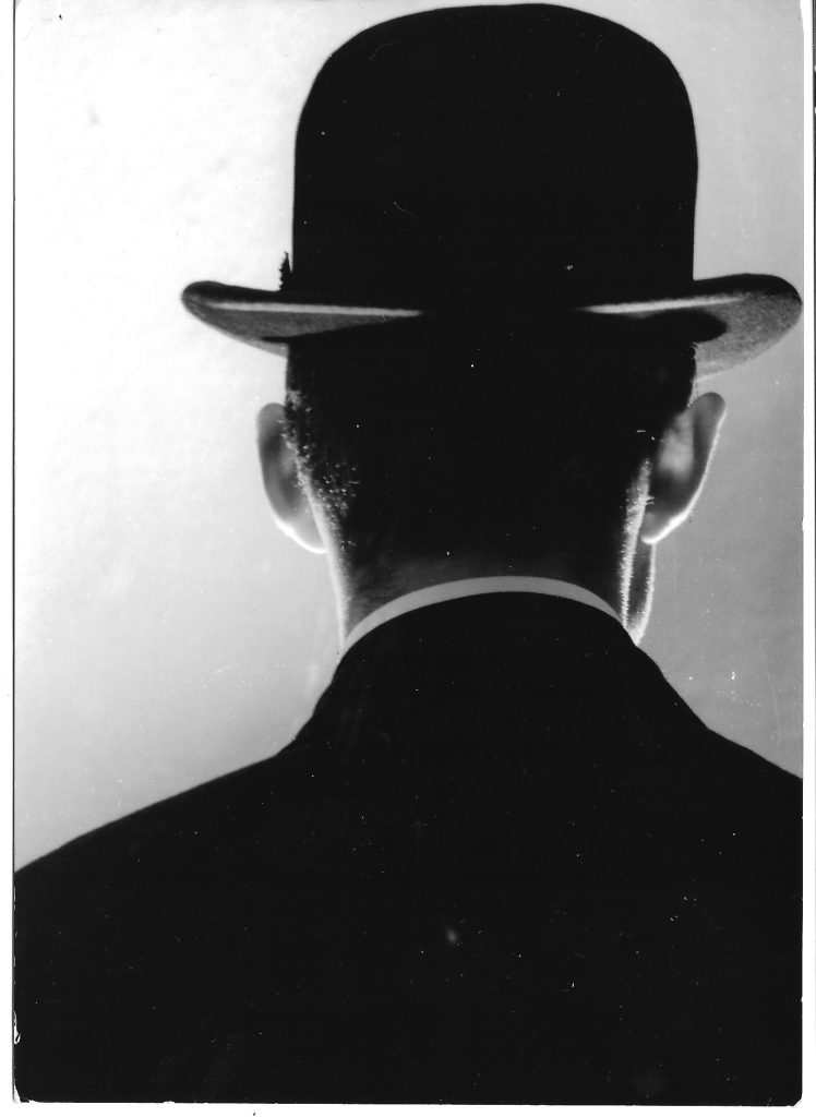 The back of a man's head. He is wearing a black suit, a white collared shirt and a black bowler hat.