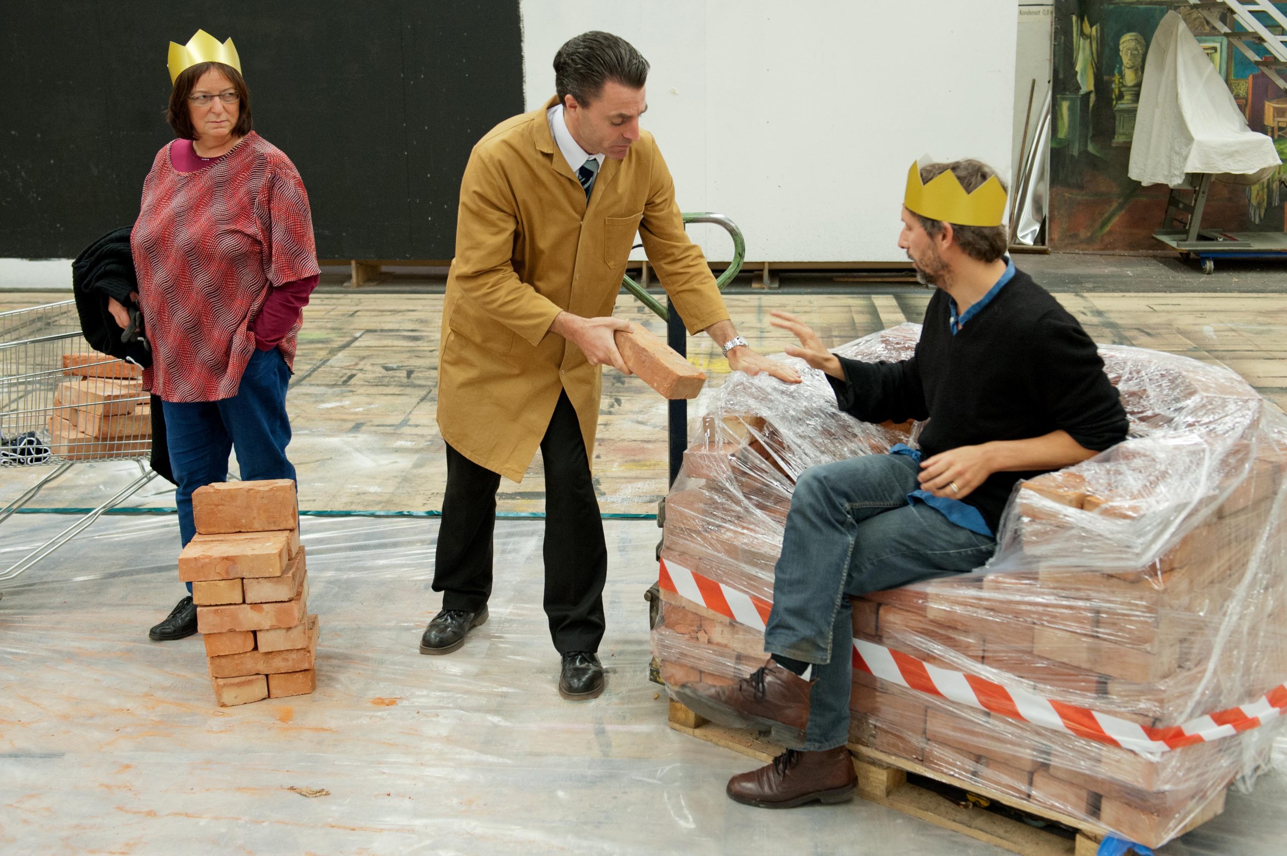 A warehouse man takes bricks from one thrown to add to a modest pile of bricks belonging to another member of the public wearing a crown.