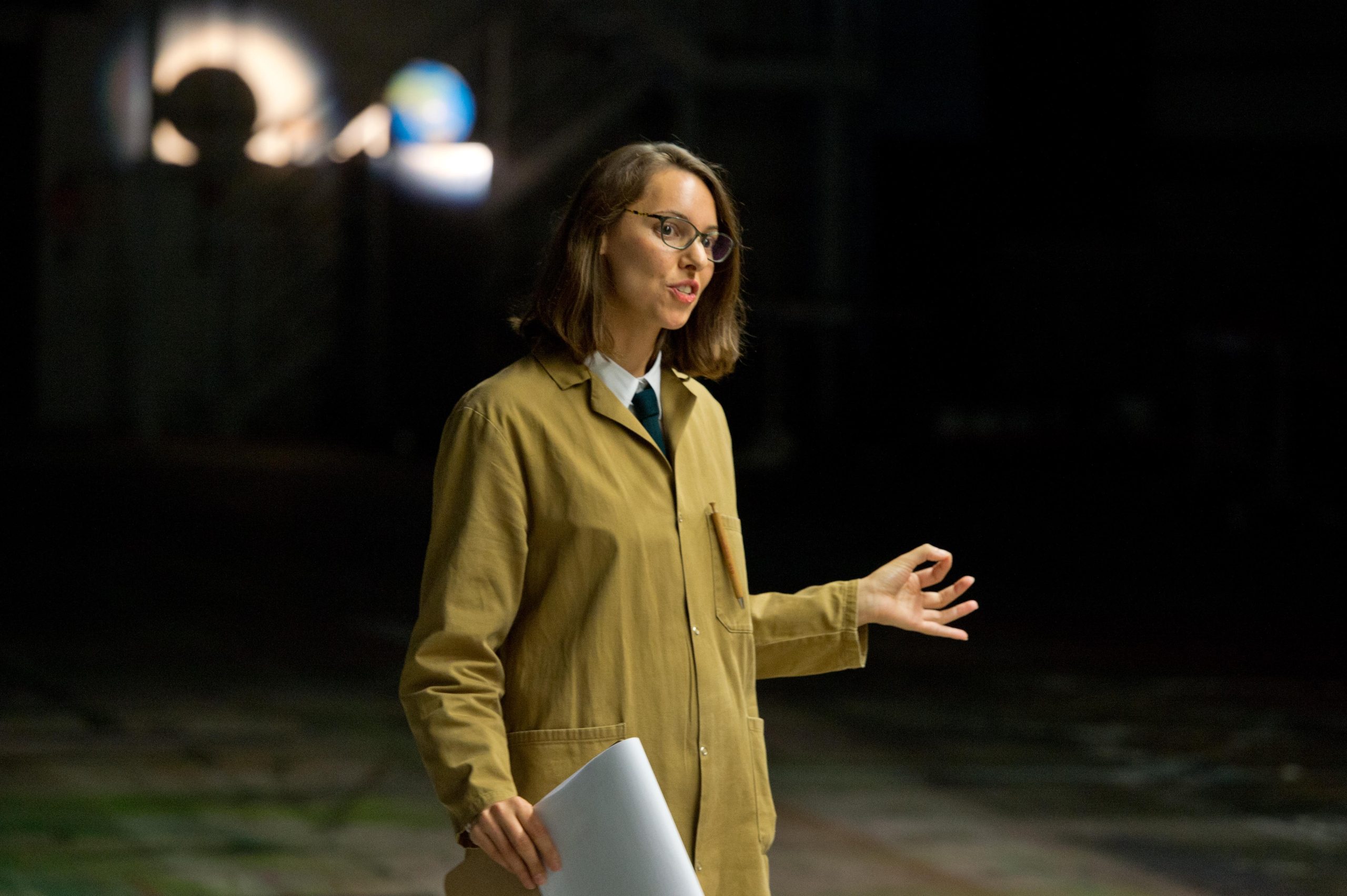 A female warehouse attendent holding a clip -board speaks, behind her in the distance, illuminated in the dark is a globe, a model of the planet earth.