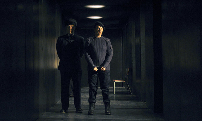 Jan Pearson in a narrow black corridor dressed in airforce blue fatigues and beret is handcuffed, a military policeman stands behid her right shoulder.
