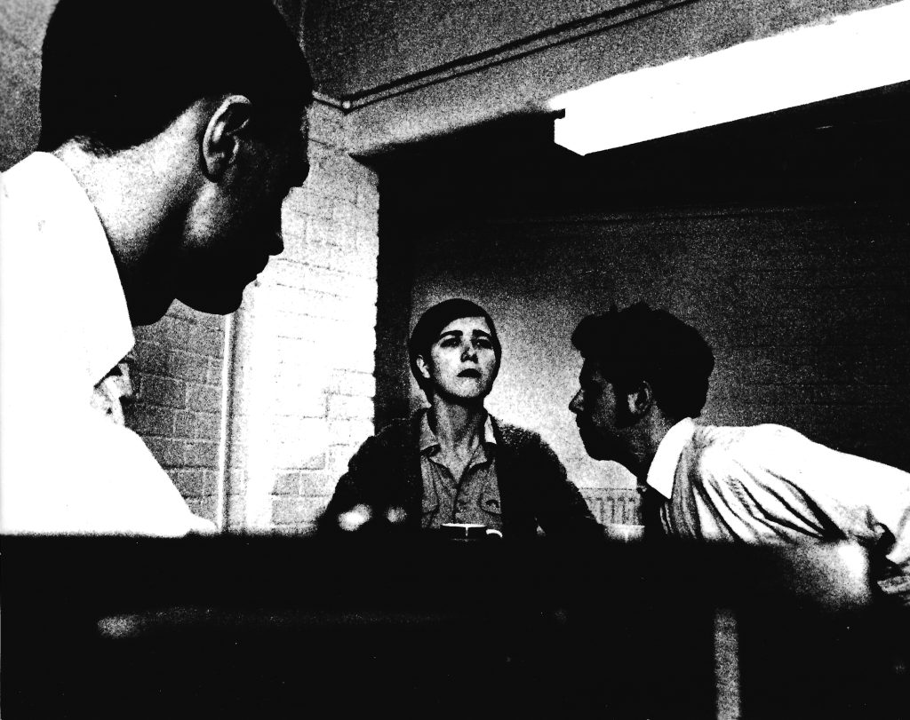 A grainy black and white image of an interrogation room. Sarah sits impassive as Nick leans into her. James sits with is back to the camera at the near end of the table.