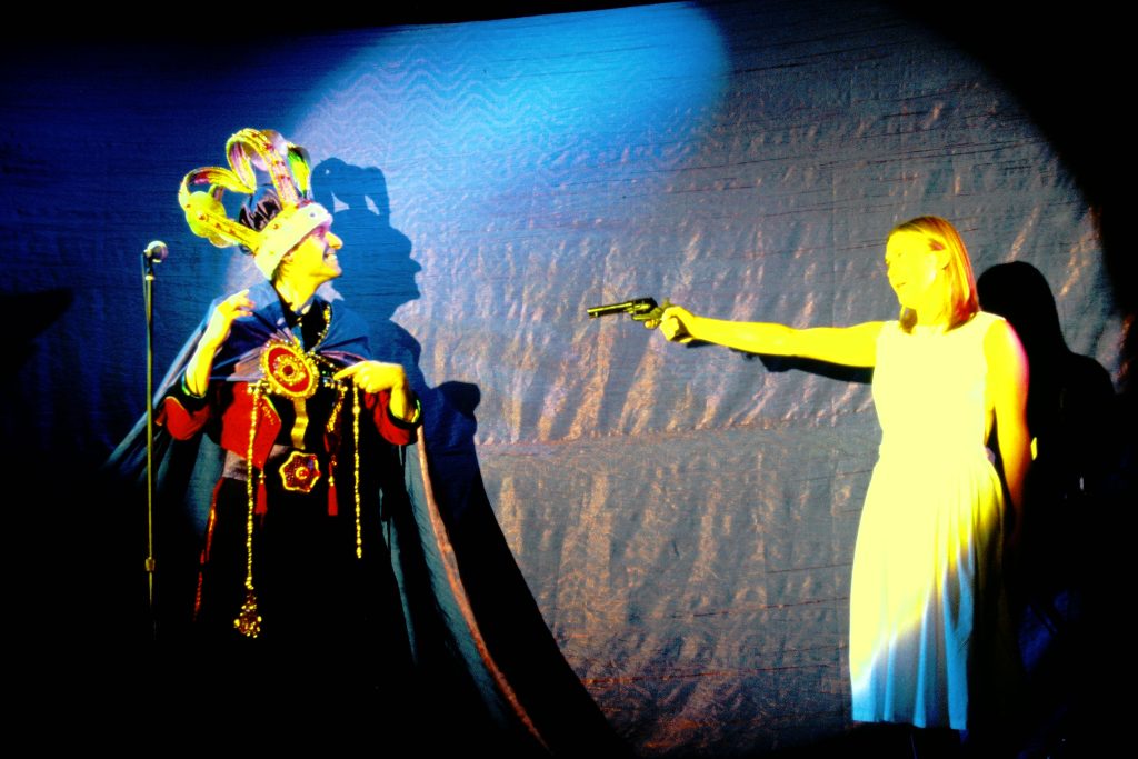 The king in an outsized crown and cape that forms a curtain obscuring the stage points to his own heart encoraging is prospective bride to shoot him as part of a circus trick.