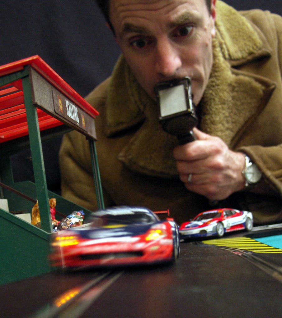 A track level shot of a man with lip mic and sheepskin coat commentating on two scalextric cars racing