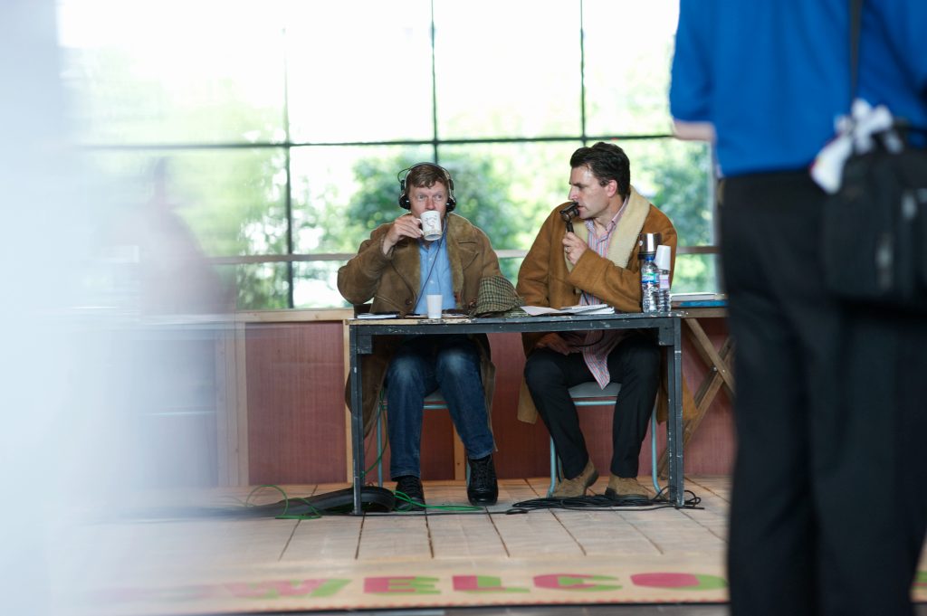 James and Craig in sheepskin coats at a table. James is commentating into a lip mic, describing Craig to his right sipping at a mug of coffee.