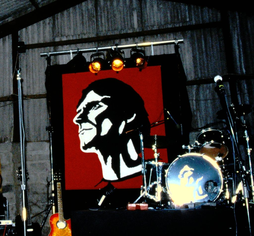 In iconic heroic rendering of Andy Watson's profile gazing clear eyed into the distance as The President on a banner behind the drum riser on stage in a barn, outside Lancaster.