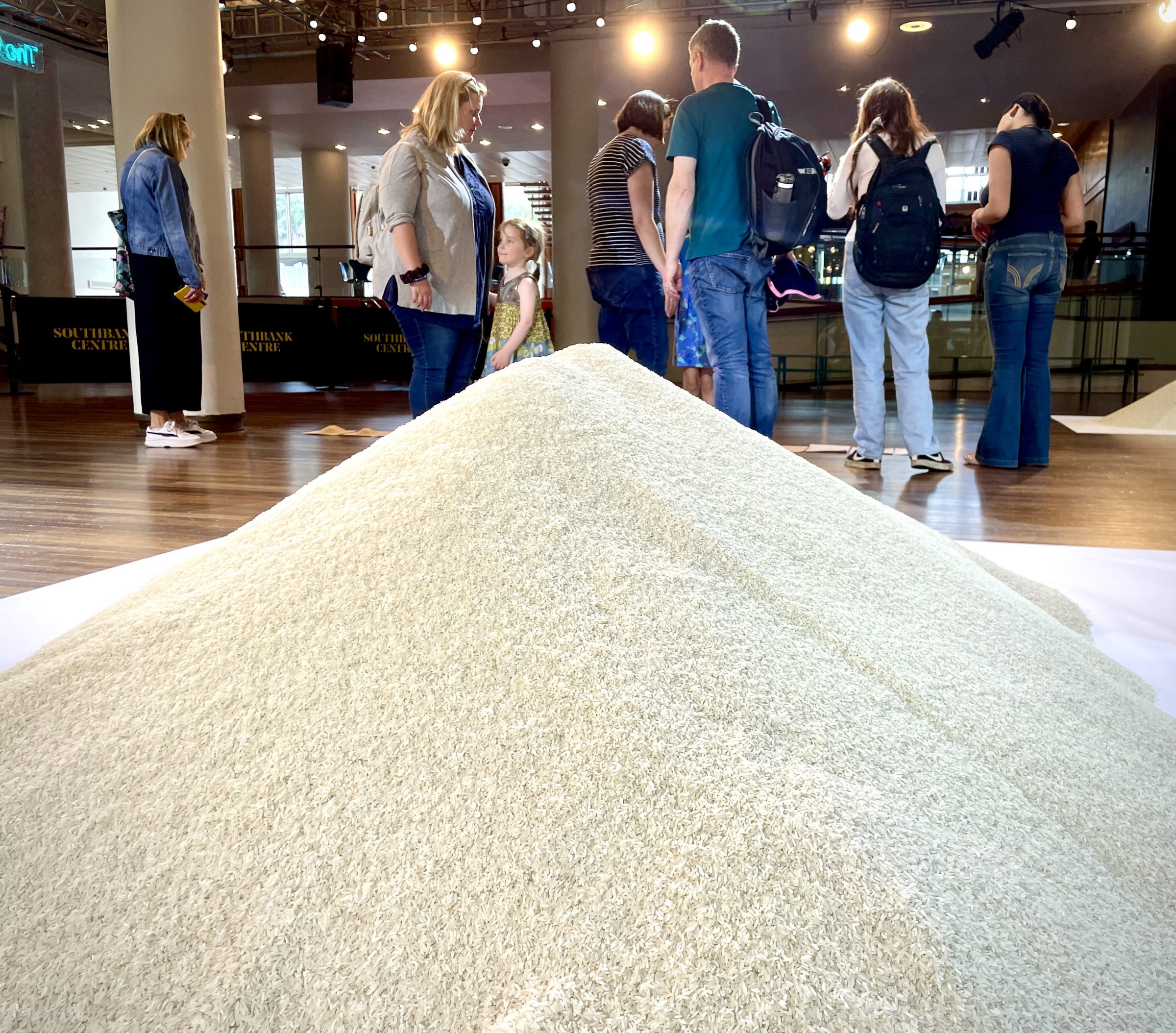 Picture of the show Of All The People In All The World at the Southbank Centre. In the foreground is a large pile of rice representing the number of refugees in the world. In the rest of the picture lots of people can be seen looking at other piles of rice.